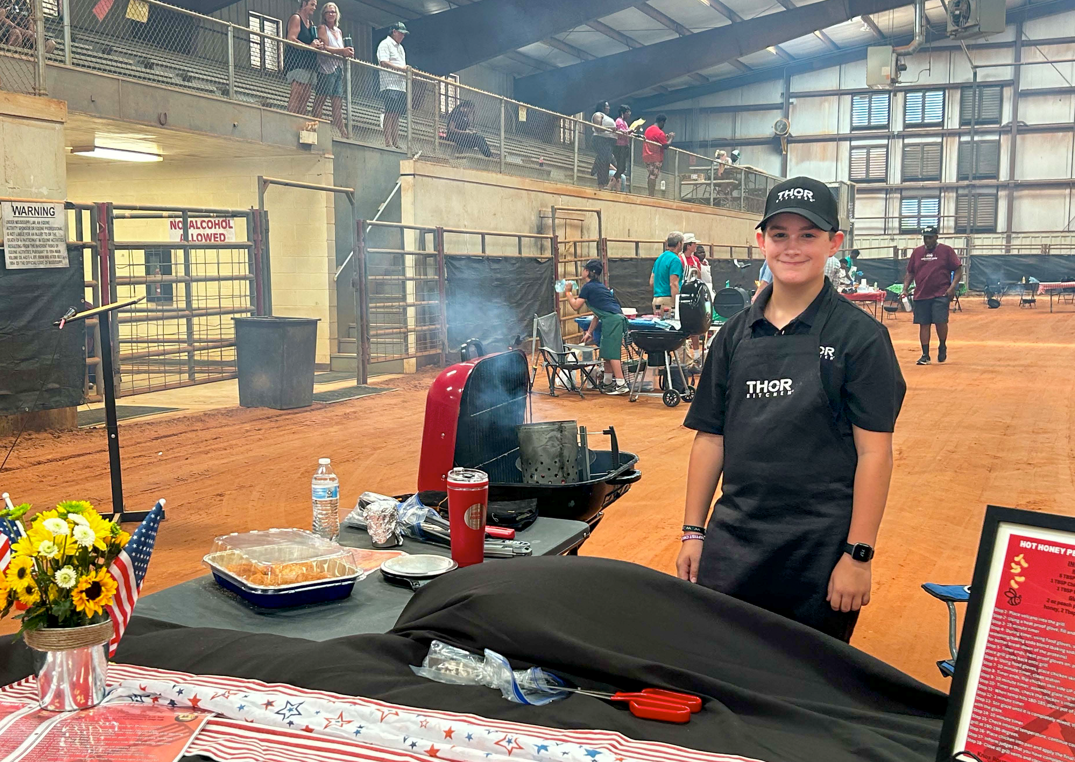 A contestant stands by a decorated table and charcoal grill.